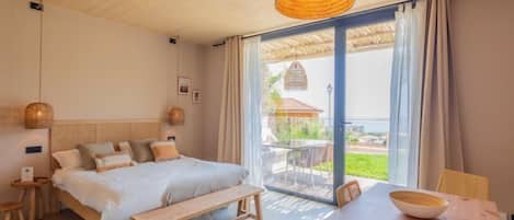 A bedroom with a bed, bedside tables, a bench, a dining table, and a view of the outdoors.