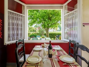 Mesa de jantar posta para quatro pessoas, com cortinas de renda, uma garrafa de vinho e vista para uma árvore verde e um jardim através de uma janela.