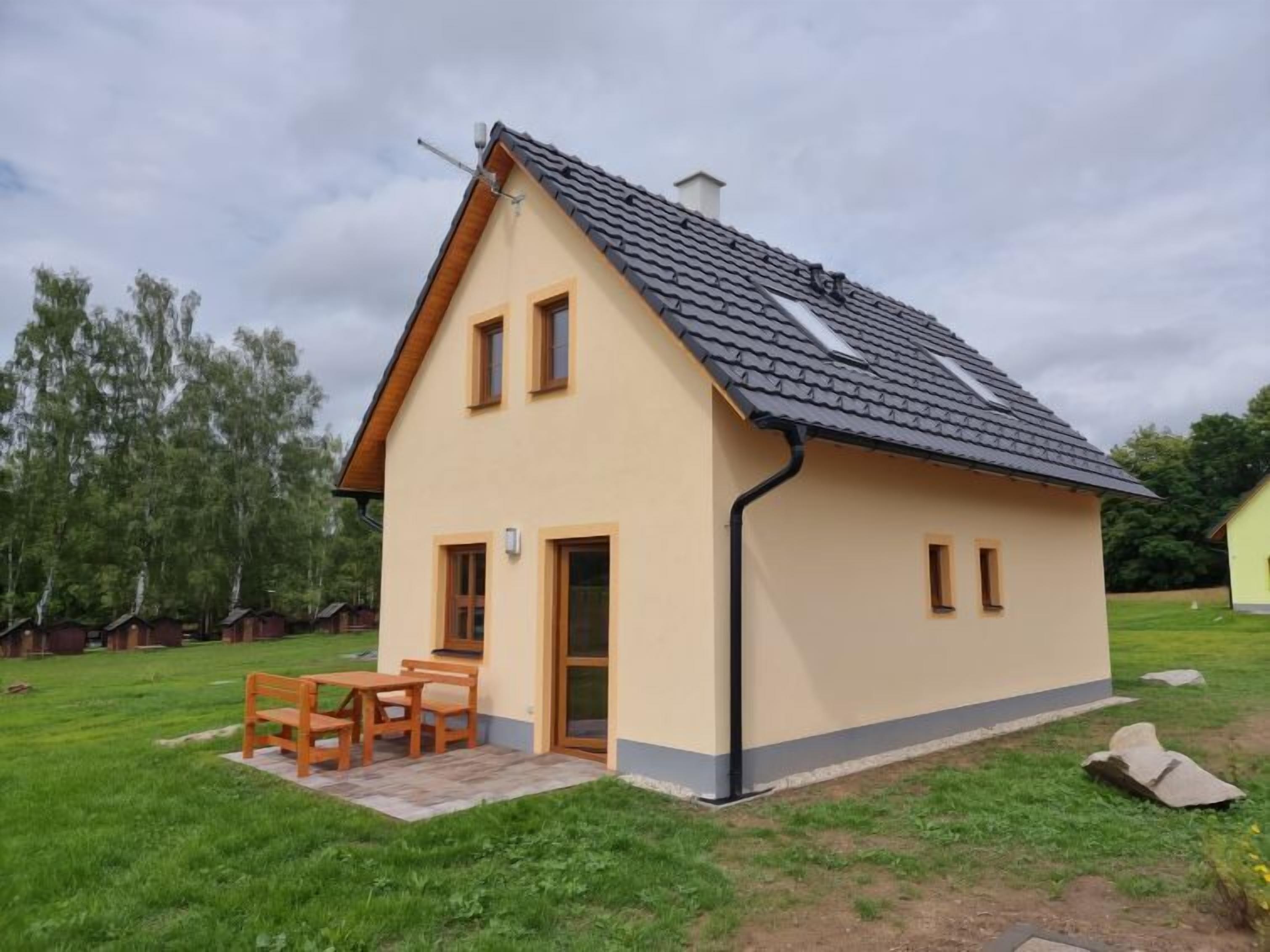 Une maison de plain-pied avec un toit en pente, un petit porche avec une table et des bancs, et un ciel dégagé.