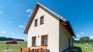 Una casa a due piani con tetto spiovente, un patio con mobili di legno e un'auto rossa parcheggiata sullo sfondo.