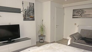 Un dormitorio moderno con un televisor de pantalla plana, un sofá gris, una colcha blanca y dos cuadros colgados en la pared.