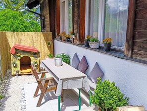 A wooden house with a small outdoor seating area, a wooden playhouse, and potted plants on the windowsill.
