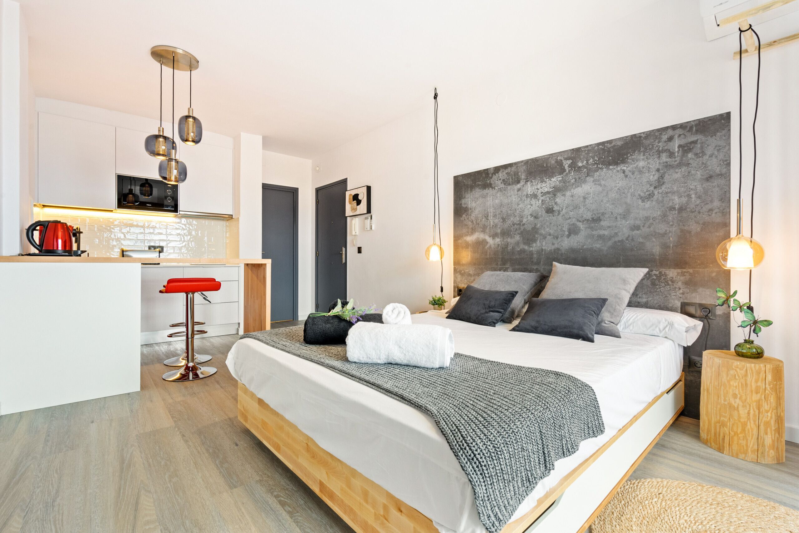 A modern hotel room with a large bed, a kitchenette, a red bar stool, and a grey textured headboard.