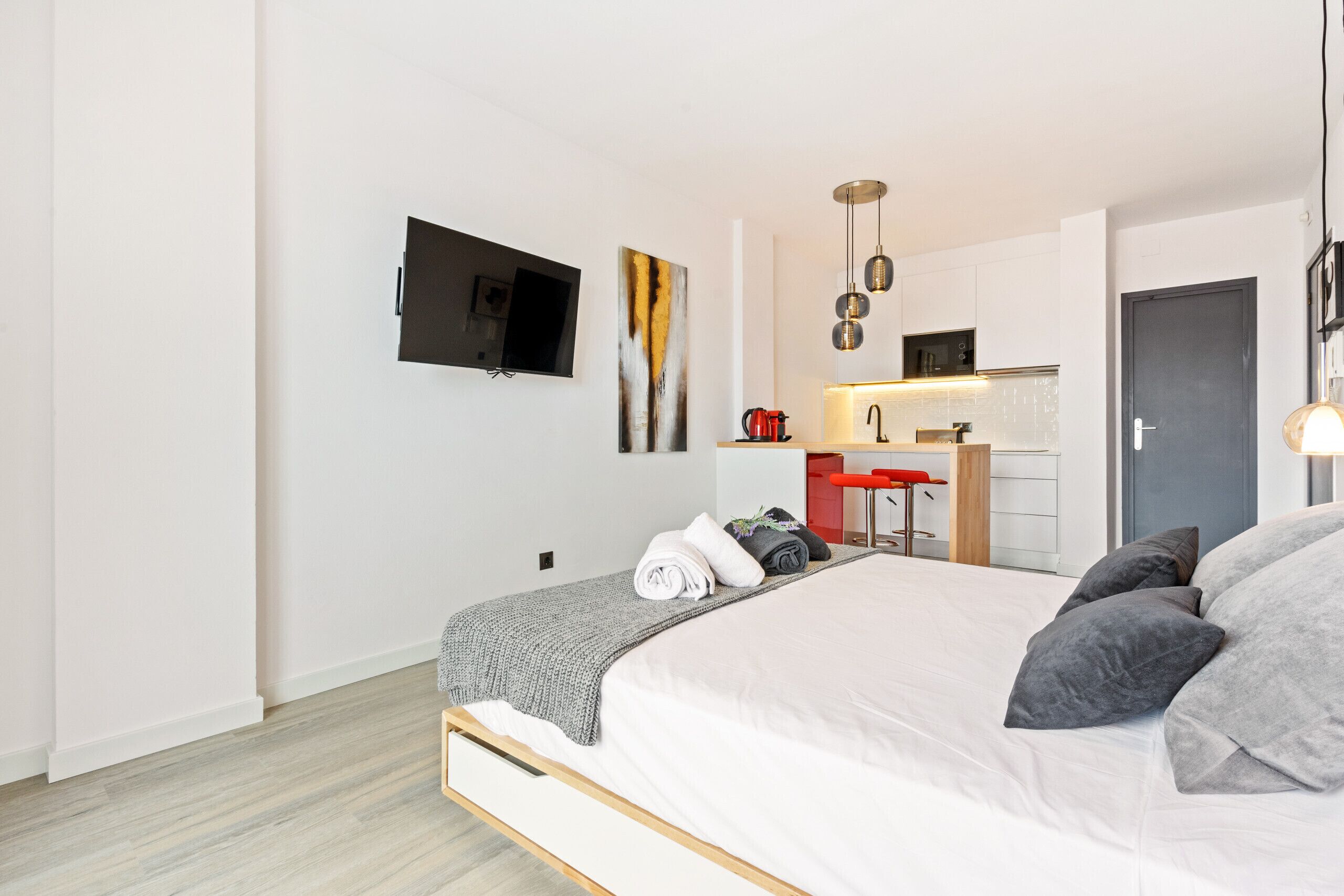 A modern bedroom with a large bed, a TV mounted on the wall, a kitchen area with a sink and countertop, and a door leading to another room.