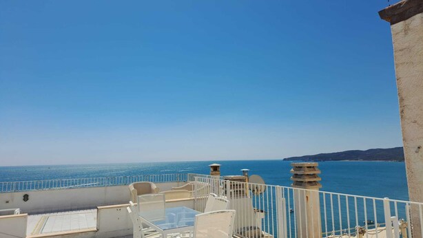 Una terraza con vista al mar, barandas blancas y un edificio de fachada de piedra.