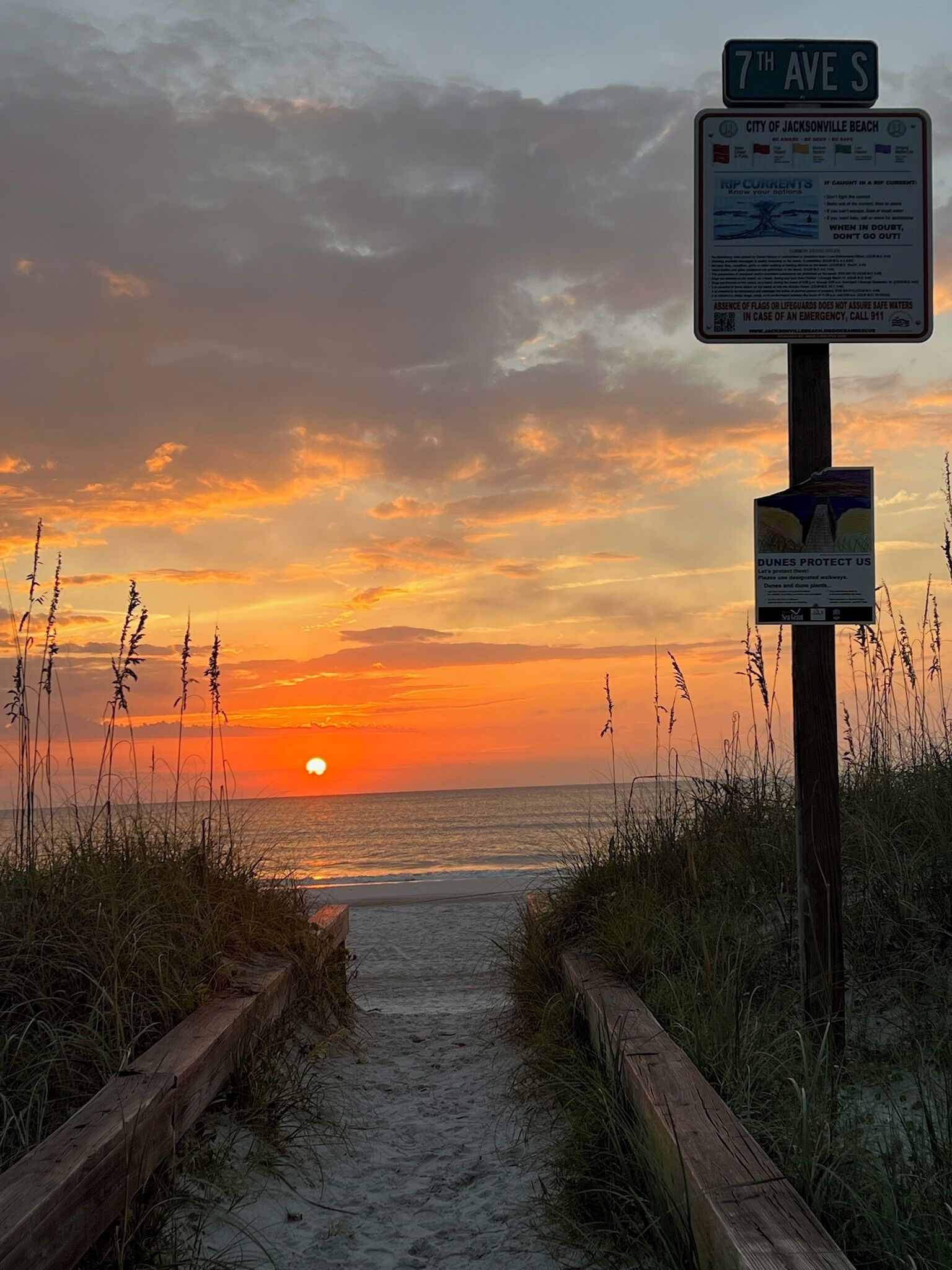 A sunset view at 7th Ave S with a wooden boardwalk leading to the beach, surrounded by tall grasses.