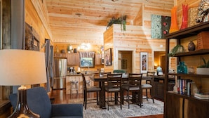 A cozy log cabin interior with a dining area, kitchen, and a bookshelf filled with books and decorative items. - Louise's Hideaway by Inn & Spa at Cedar Falls (Logan)