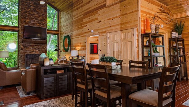 A cozy cabin interior with a dining area, wooden walls, a fireplace, and a large window overlooking greenery. - Louise's Hideaway by Inn & Spa at Cedar Falls (Logan)