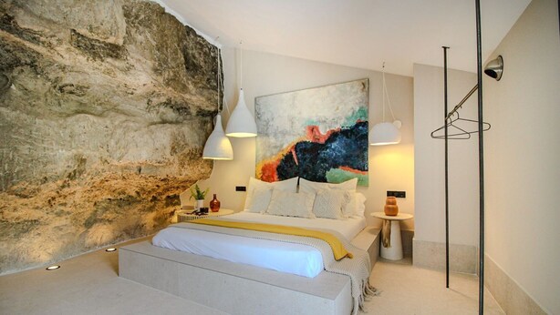 A bedroom with a stone wall, a bed with white bedding, a colorful abstract painting, pendant lights, and a floor lamp. - Cave Suite COVA (Cave Hotel Lomas de Ruvira) (Jorquera)