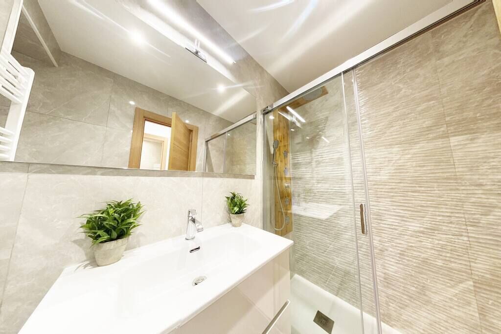 Un baño moderno con un lavamanos grande de color blanco, dos plantas en macetas, un box de ducha de cristal y una puerta de madera.