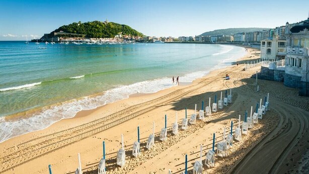 A beach with clear blue water, a sandy shore, and a hill with buildings in the background. - The Paradise: Luxurious flat, parking, jacuzzi, terrace, new (Donostia)