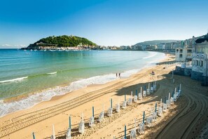 A beach with clear blue water, a sandy shore, and a hill with buildings in the background. - Paradise: Luxury apartment, parking space, Jacuzzi, terrace, new (Donostia)
