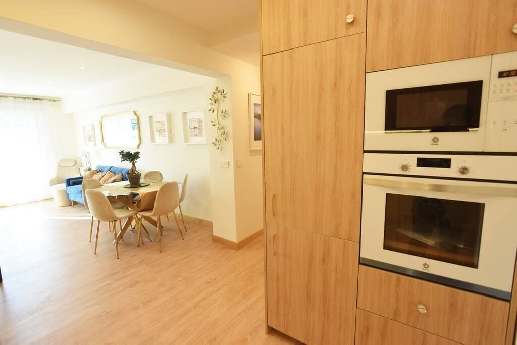 Una cocina moderna con armarios de madera y electrodomésticos integrados, un comedor con mesa y sillas, y una sala de estar con sofá y planta.