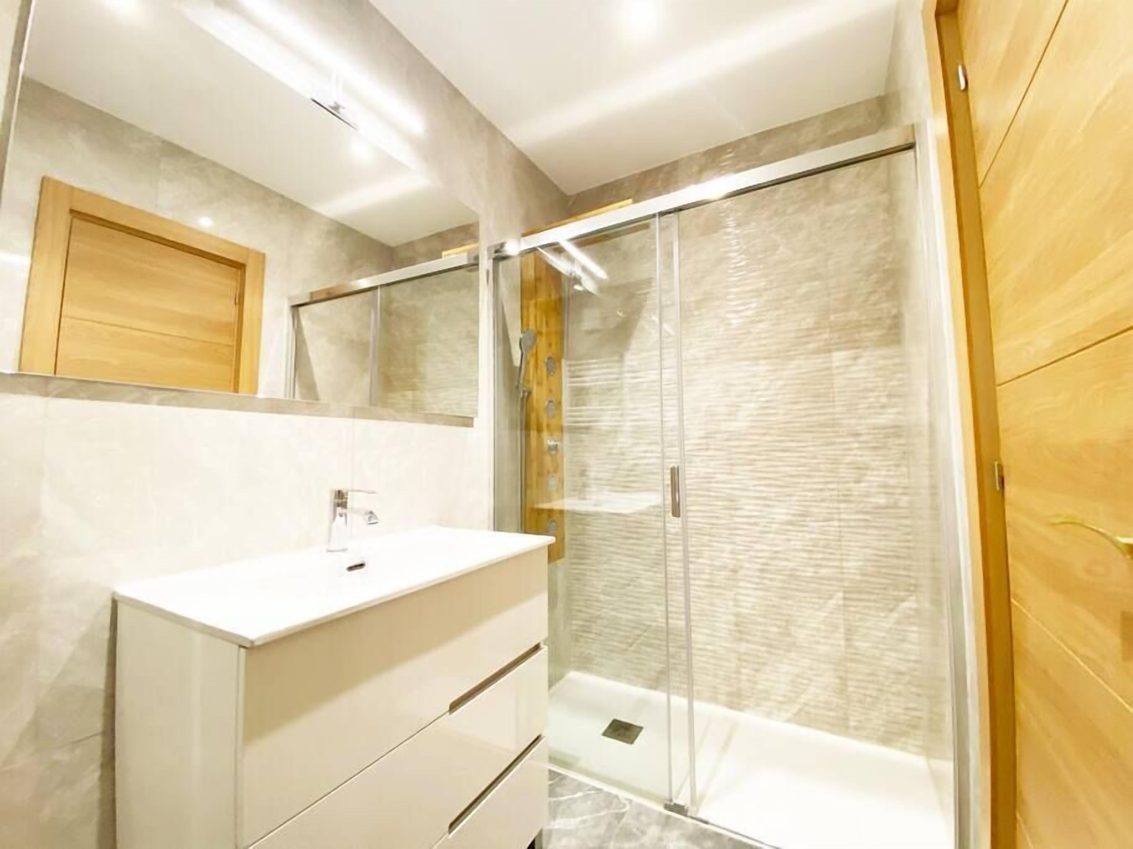 Un baño moderno con cabina de ducha de vidrio, tocador blanco y puerta de madera.
