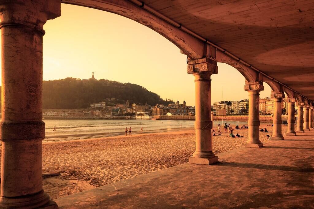 Una playa con un paseo techado sostenido por columnas, que da al mar y una colina con edificios al fondo.