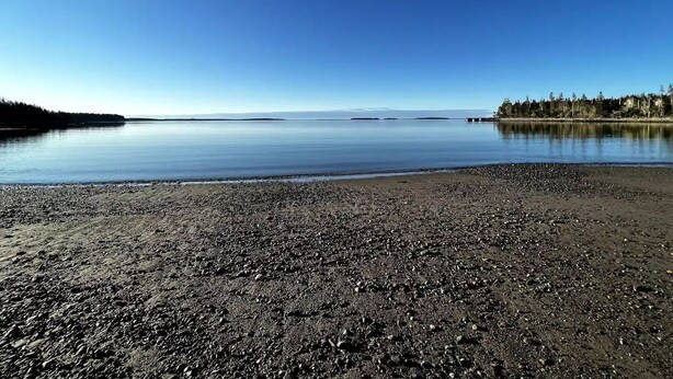A calm lake with a pebbly shore and a forested island in the distance.