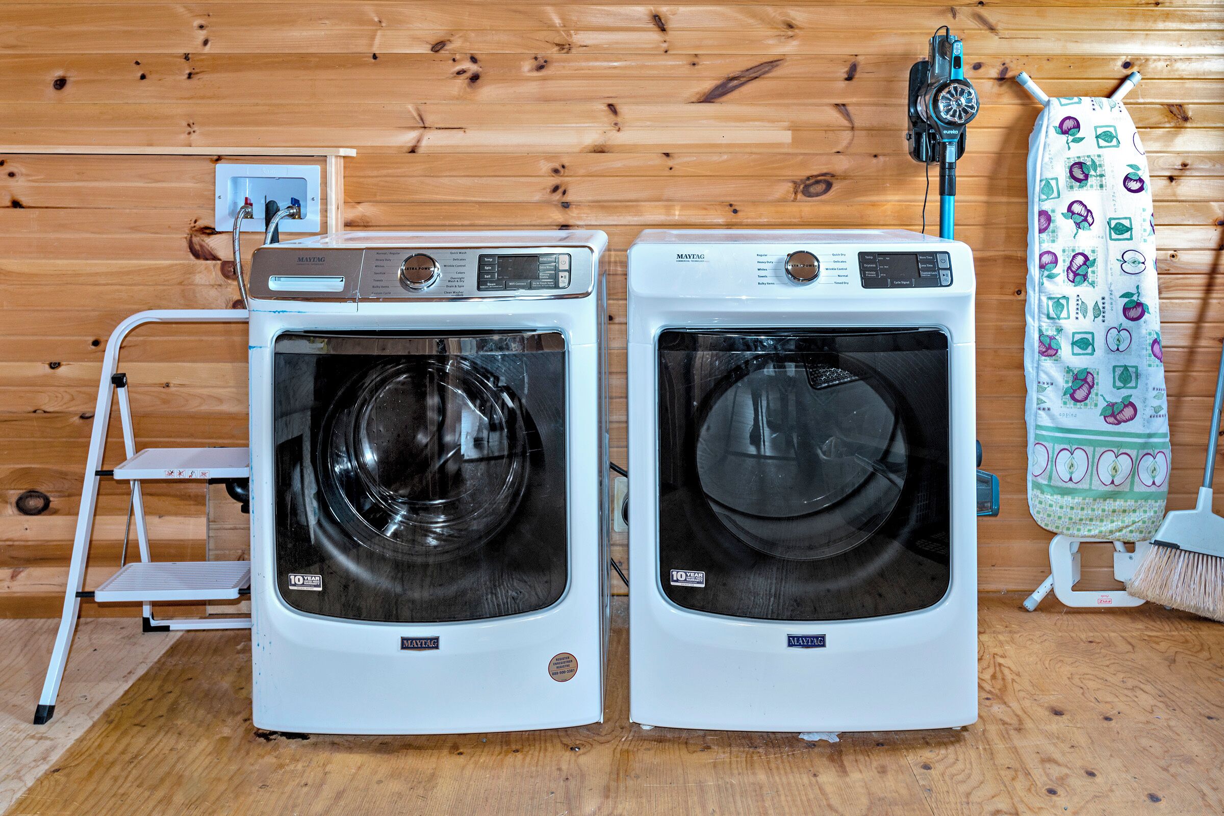 Two front-loading washing machines in a wooden room with a step ladder and a mop.