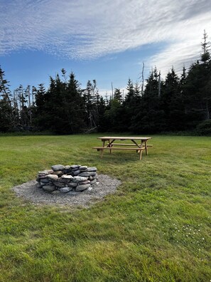 Una mesa de picnic y un hogar de piedra en un campo cubierto de césped, con árboles al fondo.