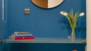 Ett rum med en blå vägg, en rund spegel, ett skrivbord i glas, en röd stol och en vas med vita blommor.