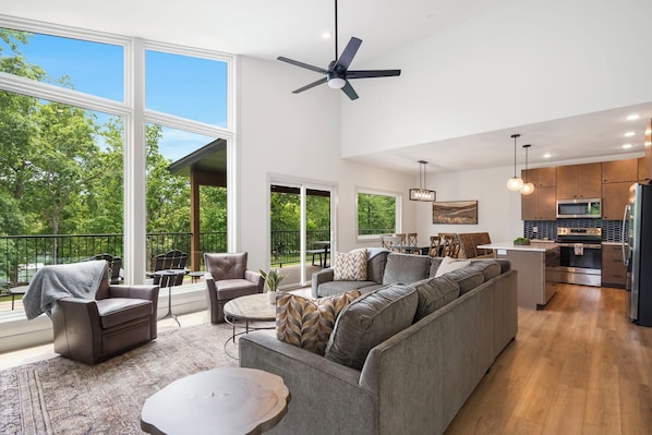 A spacious living area with a large gray sofa, a coffee table, and a ceiling fan. There is a kitchen area with modern appliances and a dining table. Large windows provide a view of the outdoors.