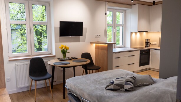 Quarto moderno com cama, mesa de cabeceira e televisão. Há uma pequena área de jantar com mesa e cadeiras, e uma cozinha com fogão e pia.