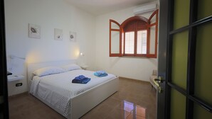 Un dormitorio con cama, mesitas de noche, ventana con contraventanas de madera y aire acondicionado de pared.