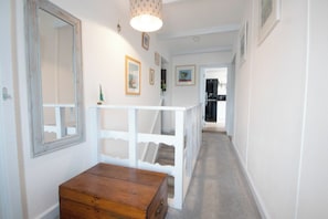Un couloir avec un banc en bois, des rampes blanches, des tableaux encadrés et un escalier menant à une cuisine.