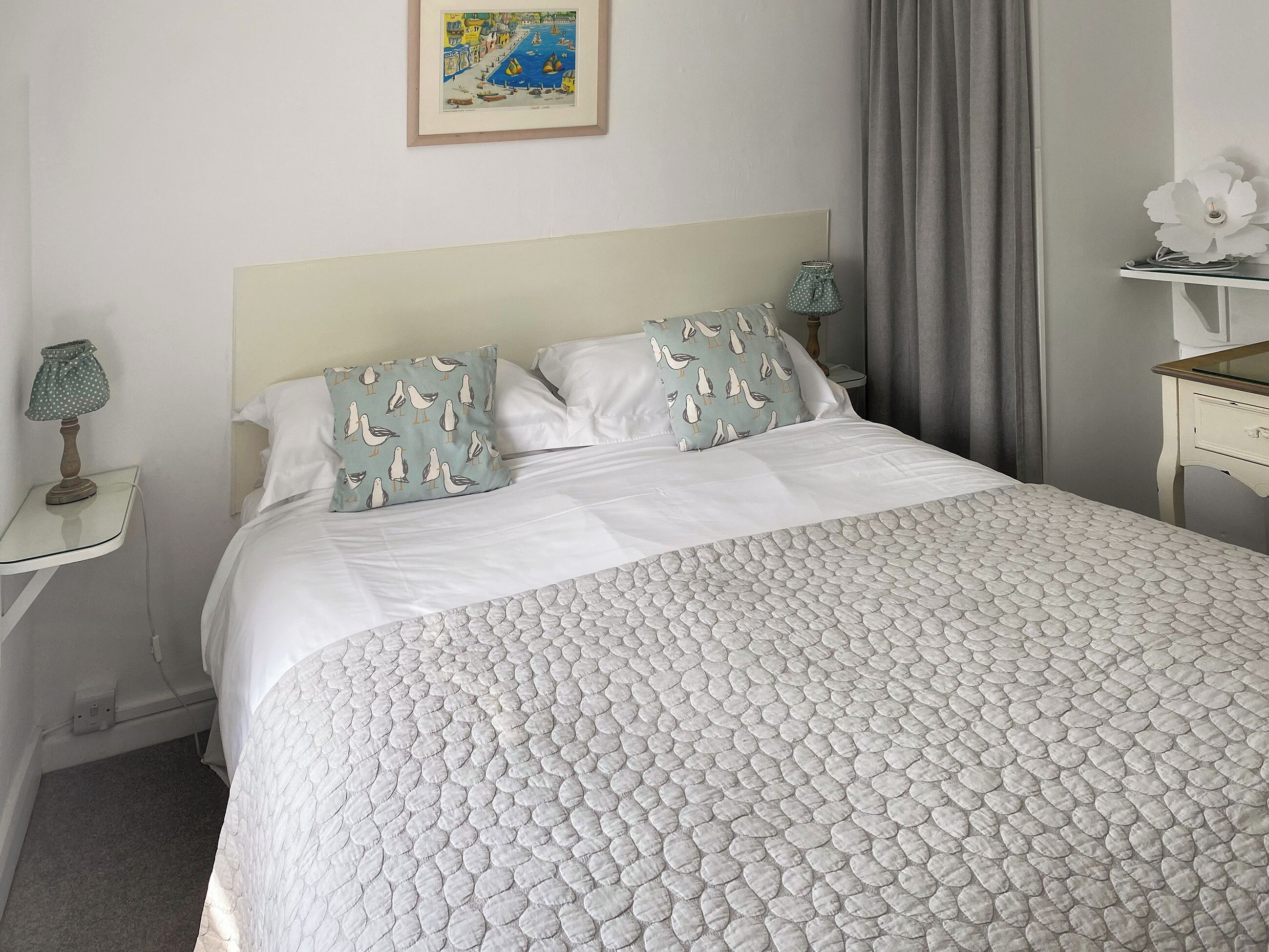 Una cama bien hecha con colcha estampada, dos almohadas con estampado de gaviotas, un cuadro en la pared, una lámpara de noche y una mesita con una flor.