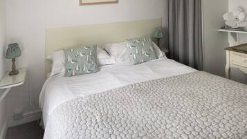 Una cama bien hecha con colcha estampada, dos almohadas con estampado de gaviotas, un cuadro en la pared, una lámpara de noche y una mesita con una flor.
