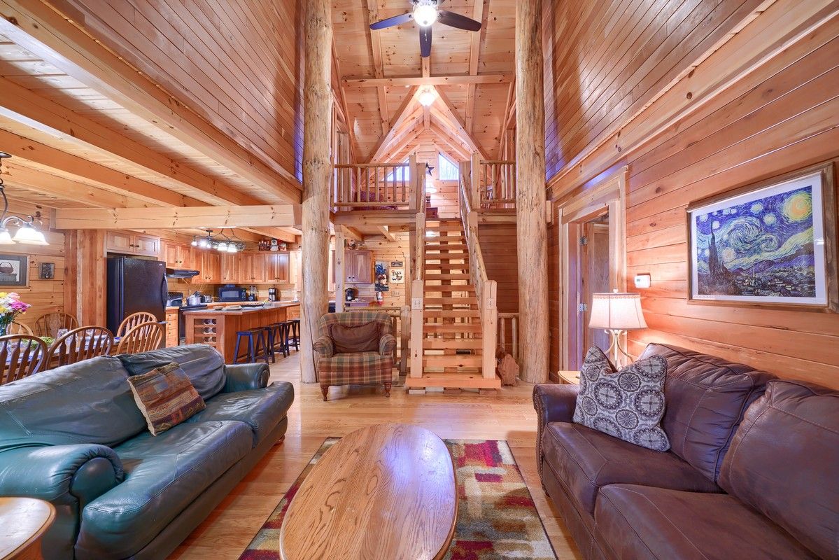 A wooden cabin interior with a dining area, two sofas, a coffee table, and a staircase leading to an upper level.