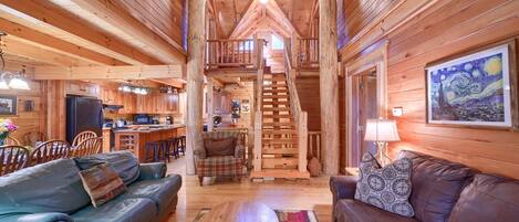 A wooden cabin interior with a dining area, two sofas, a coffee table, and a staircase leading to an upper level.