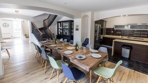 Une salle à manger spacieuse, avec une longue table en bois dressée pour six personnes, entourée de chaises aux couleurs vives. Un escalier avec une rampe en bois mène à l’étage supérieur.