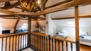 Une pièce de style « loft » avec des poutres en bois apparentes, un escalier doté d’une balustrade noire et un lit en mezzanine avec une tête de lit bleue.