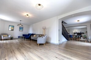 Een ruime woonkamer met houten vloer, een blauwe bank, een grijze fauteuil en een eethoek met een trap.