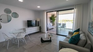 Una sala de estar moderna con una mesa de comedor de vidrio, sillas blancas, un sofá con cojines de colores, un televisor de pantalla plana y un ventanal corredizo que da a un área exterior.