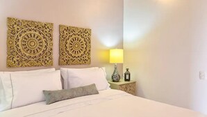 A neatly made bed with white linens and a single decorative pillow, flanked by two ornate wall decorations. A bedside table with a lamp and a small vase is visible. - 2CM-B1 APARTAMENTO EN GETSEMANI CON BALC (Cartagena)