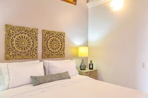 A neatly made bed with white linens and a single decorative pillow, flanked by two ornate wall decorations. A bedside table with a lamp and a small vase is visible. - 2CM-B1 APARTAMENTO EN GETSEMANI CON BALC (Cartagena)