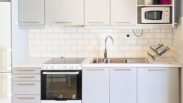 Una cucina compatta con mobili bianchi, forno integrato, lavello in acciaio inossidabile e forno a microonde su un ripiano.
