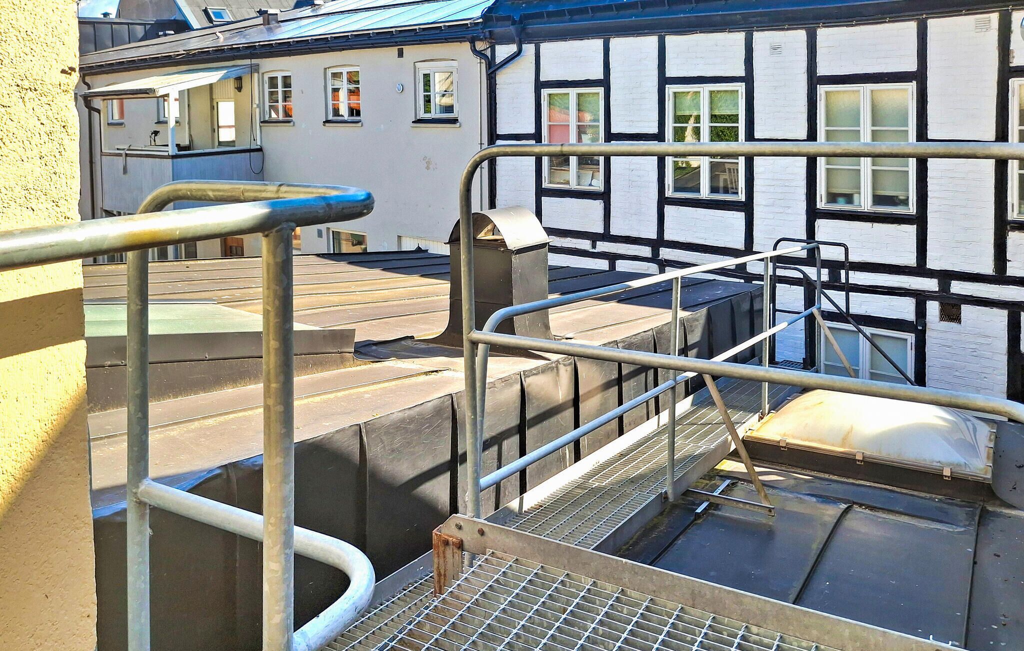Eine Metalltreppe führt zu einer Dachterrasse mit Metallgeländer und Metallgitterrost. Im Hintergrund ein Gebäude mit weiß-schwarzen Verzierungen.
