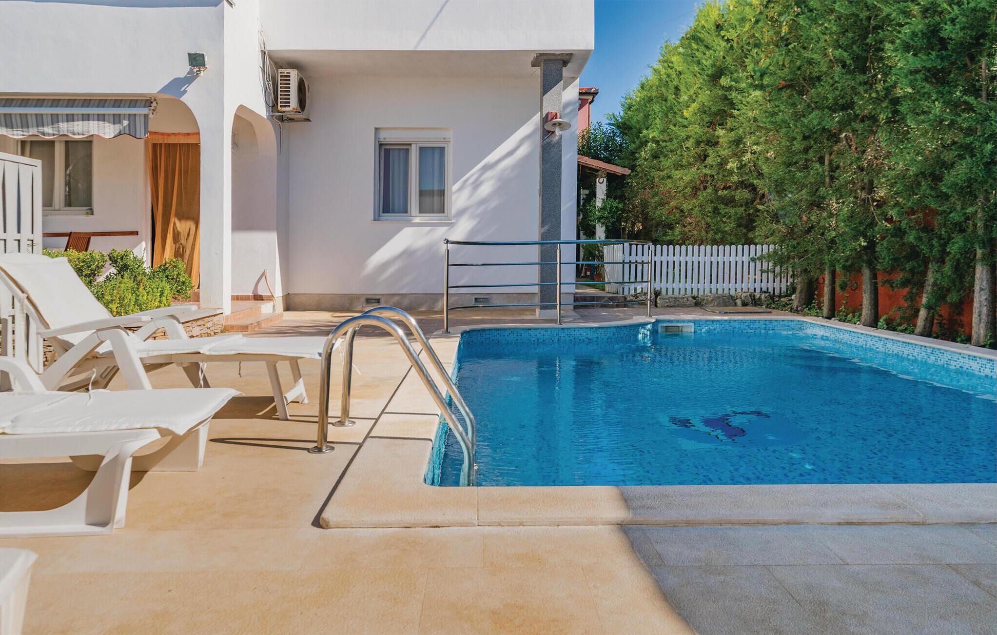 A swimming pool with a metal ladder, white lounge chairs, and a white building with a window and air conditioning unit.
