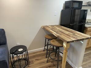 A compact kitchen with a wooden island, two stools, a black round table, and a microwave above the refrigerator.