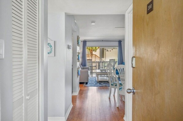 A hallway leading to a dining area with a table and chairs, a wooden door with a number sign, and a view of the outside. - Oceanfront 2BR • Pool • Walk Beach • Sleeps 6 (Fernandina Beach)