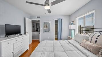 Un dormitorio con cama, televisor, cómoda, ventilador de techo y ventana con persianas.