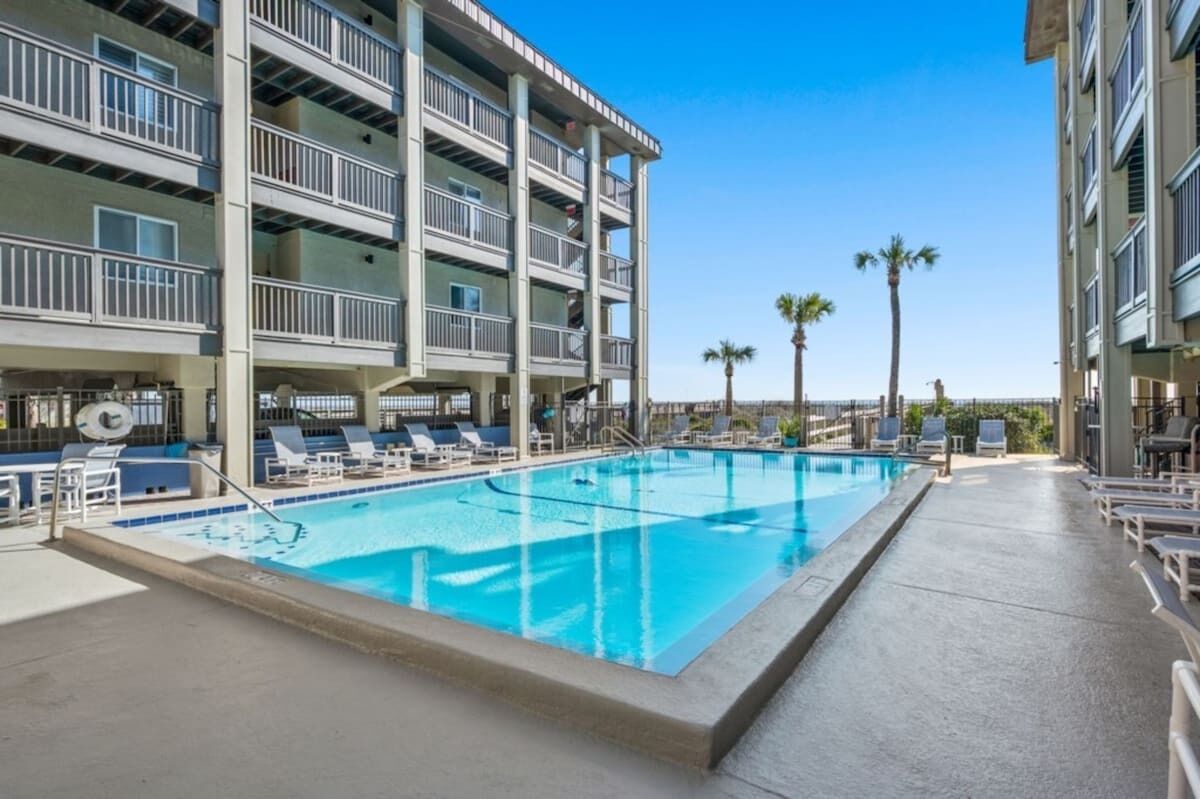 Una piscina con sillas de descanso, un edificio de varios pisos con balcones y palmeras.