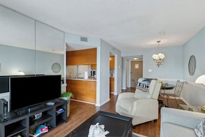 A living room with a TV, sofa, and coffee table. There is a kitchen in the background. - Beachfront 2BR • Pool • Direct Access (Fernandina Beach)