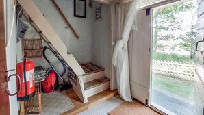 Una habitación pequeña y acogedora con una escalera que sube a una zona superior, una ventana con vistas al verdor, y un extintor en el suelo.