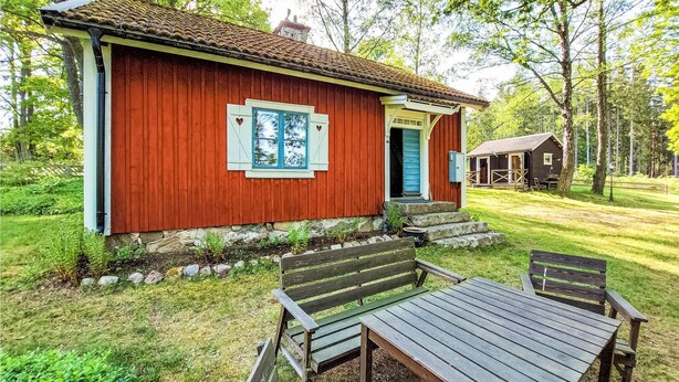 Una cabaña de madera roja con puerta y ventanas azules, un pequeño porche y un juego de mesa y banco afuera.