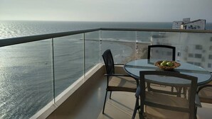 Un balcó amb barana de vidre que dona al mar, una taula rodona amb cadires i una cistella de fruita.