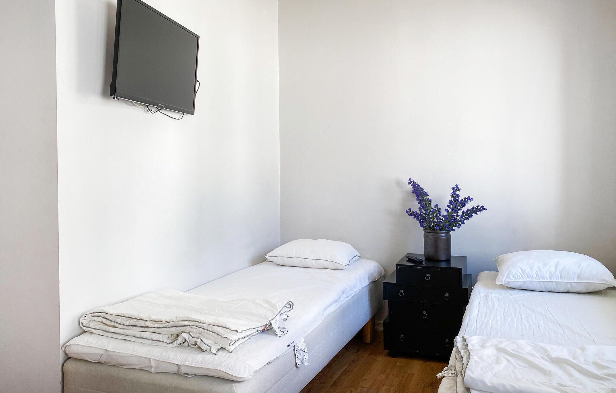 A small, clean room with two single beds, a flat-screen TV mounted on the wall, and a small table with a vase of flowers.