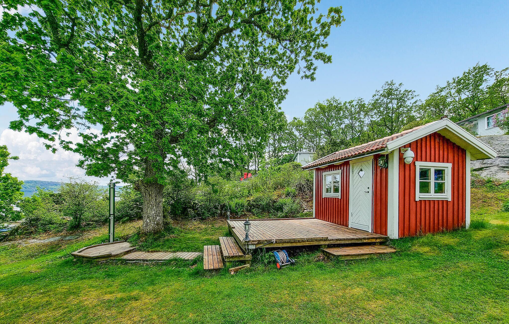 Une cabane rouge avec une terrasse en bois et une porte blanche, entourée d’une végétation luxuriante et d’arbres.